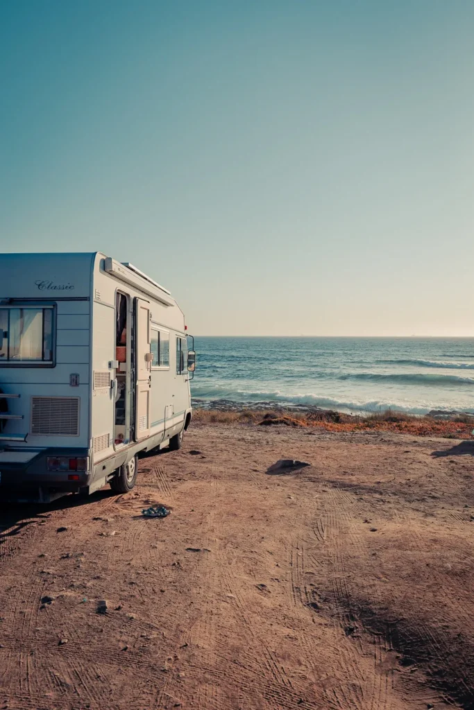 A camper van is parked on a sandy area facing the Atlantic Ocean near Praia de Oliveirinha, under a clear sky in summer.