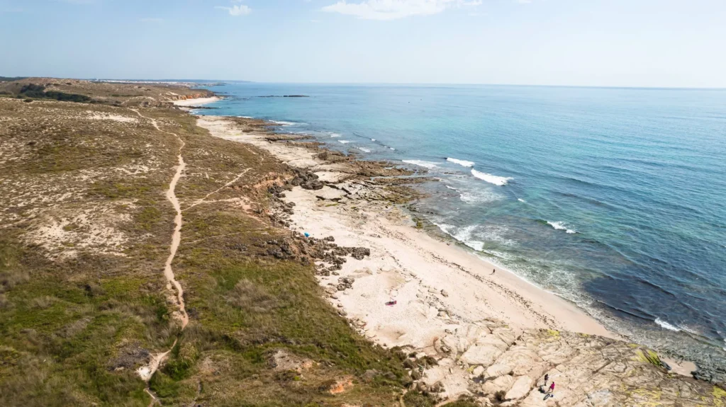 Aerial view of Praia de Oliveirinha near Porto Covo, showing a wild beach lined with rocks and the Atlantic Ocean under a clear sky in June.
