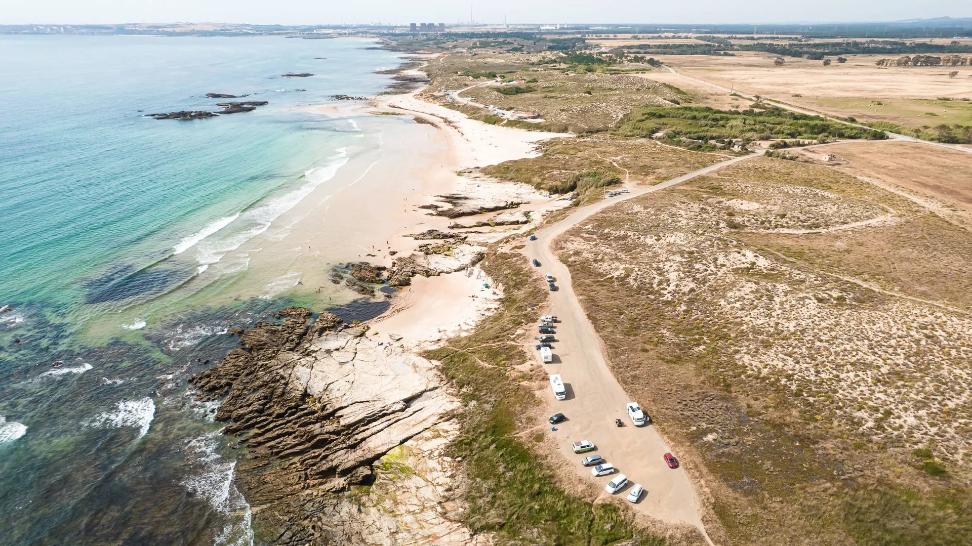 Aerial view of Oliveirinha beach near Porto Covo, in Alentejo, Portugal, showing a vast stretch of sand, seaside rocks, and a parking lot with a few cars in June 2026.