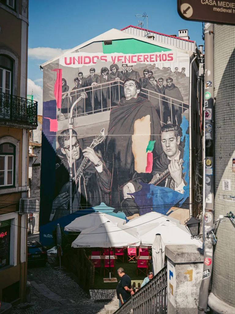 Mural depicting musicians and a choir with the inscription 'Unidos Venceremos', in a cobbled alley of Coimbra under a clear sky, with a café terrace in the foreground.