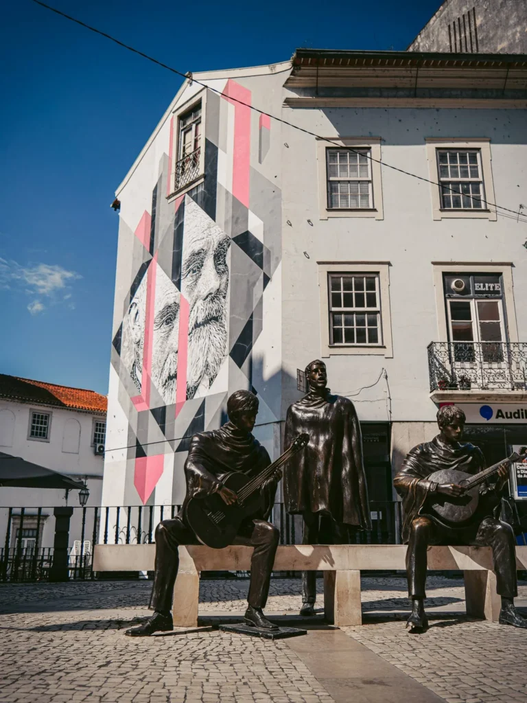 Musician statues of fado on a cobbled square in Coimbra, in front of a mural depicting a bearded face on the facade of a building.