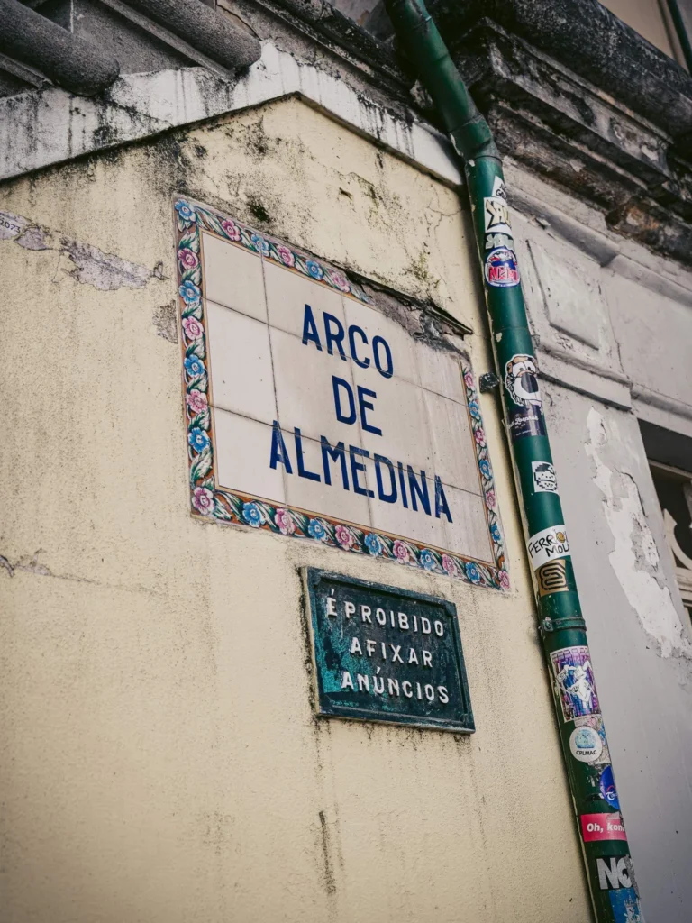 Tile plaque indicating 'Arco de Almedina' on an old wall in Coimbra, Portugal, with a pipe adorned with numerous stickers.