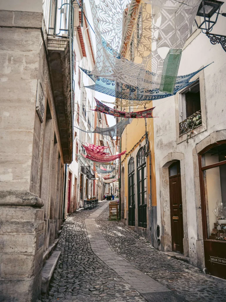 Narrow cobbled street in the historic center of Coimbra decorated with colorful nets hanging between the buildings, calm atmosphere with no visible human presence.