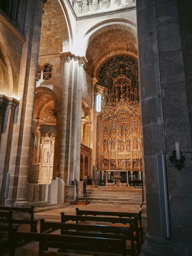 Interior of a historic church in Coimbra, featuring a richly decorated altar and stone vaults typical of Portuguese Romanesque architecture. No visitors are visible in the nave.