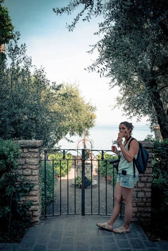 Caroline is eating an ice cream in front of a wrought iron gate surrounded by vegetation in Sirmione.