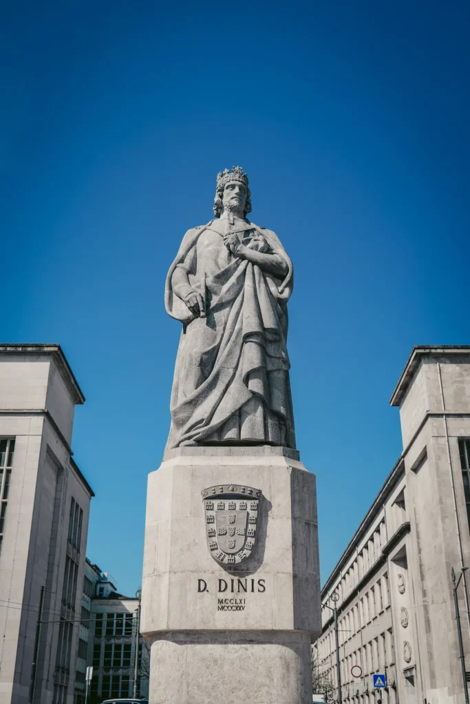 Statue of D. Dinis in a large urban square in Coimbra under a blue sky, surrounded by modern buildings.