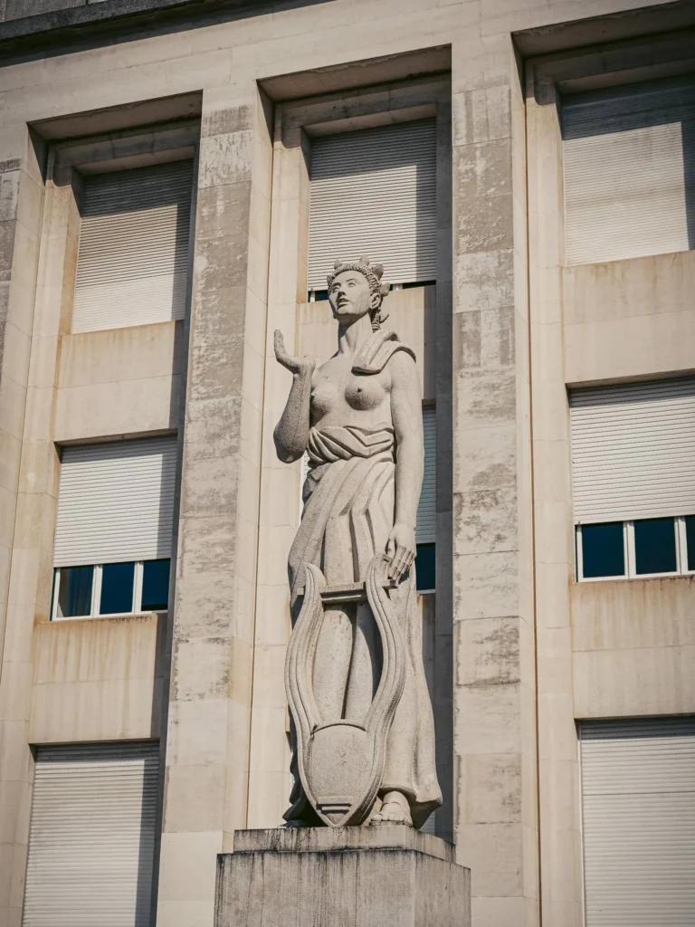 Monumental stone statue representing a woman with a lyre in front of a building facade in Coimbra, Portugal, under daylight.