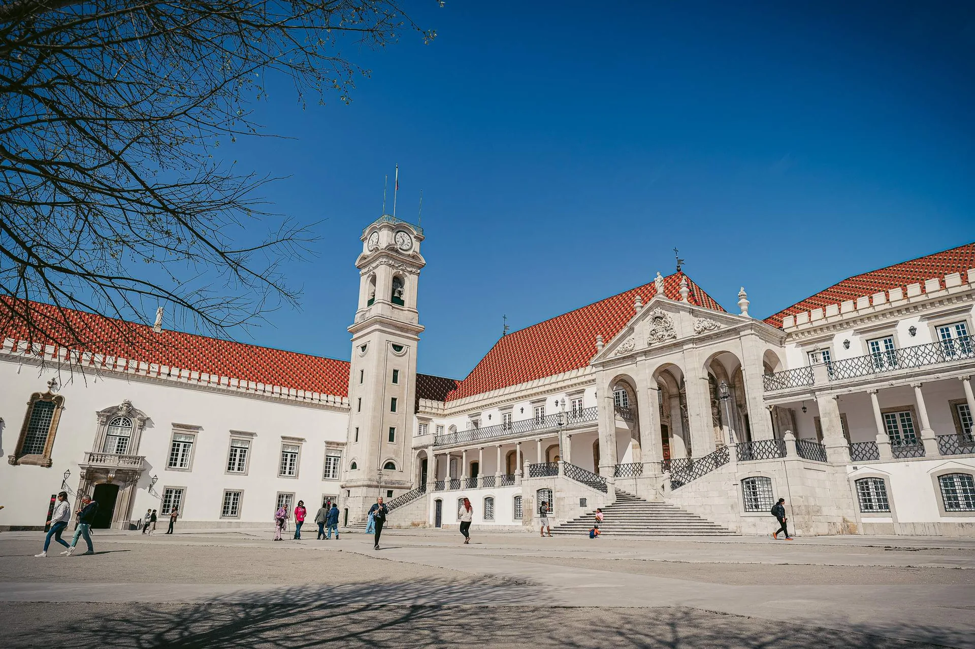 The main courtyard of the University of Coimbra in Portugal, with its famous tower and visitors strolling under a blue sky.