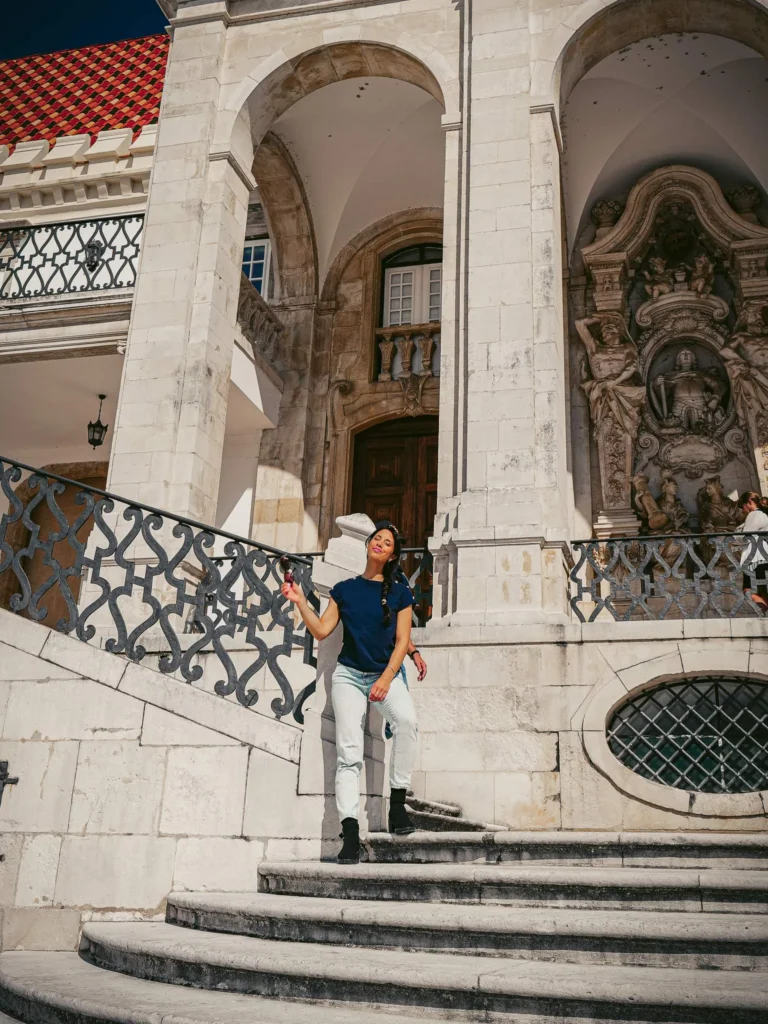 Caroline poses on the stairs of a historic building in Coimbra, under a stone arch and in front of a façade adorned with baroque sculptures.