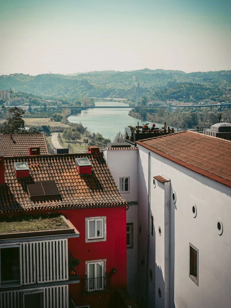 View of the rooftops of Coimbra with the Mondego River and lush hills in the background, under a clear sky.