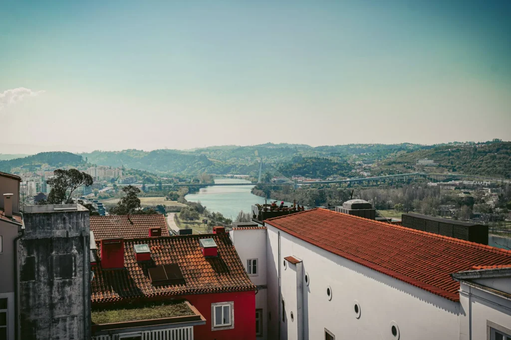 Panoramic view of the rooftops of Coimbra with the Mondego River and lush hills in the background, under a clear sky in Portugal at the end of March.
