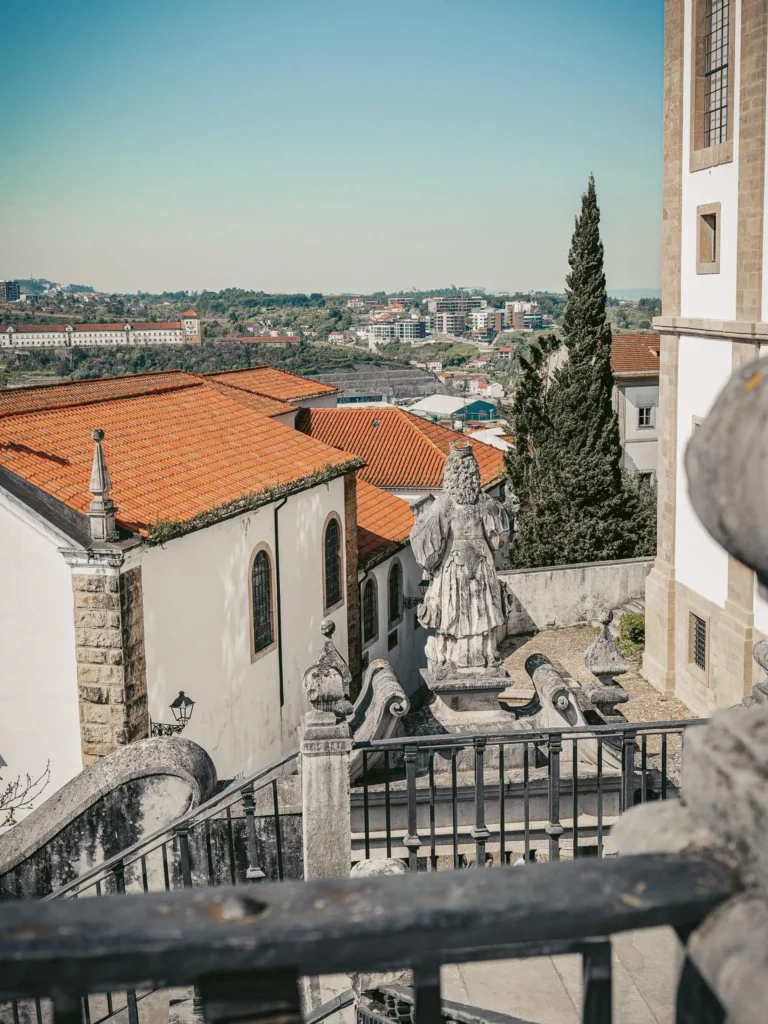 View of the red-tiled rooftops and a stone statue in Coimbra, with the city and countryside in the background under a clear sky.