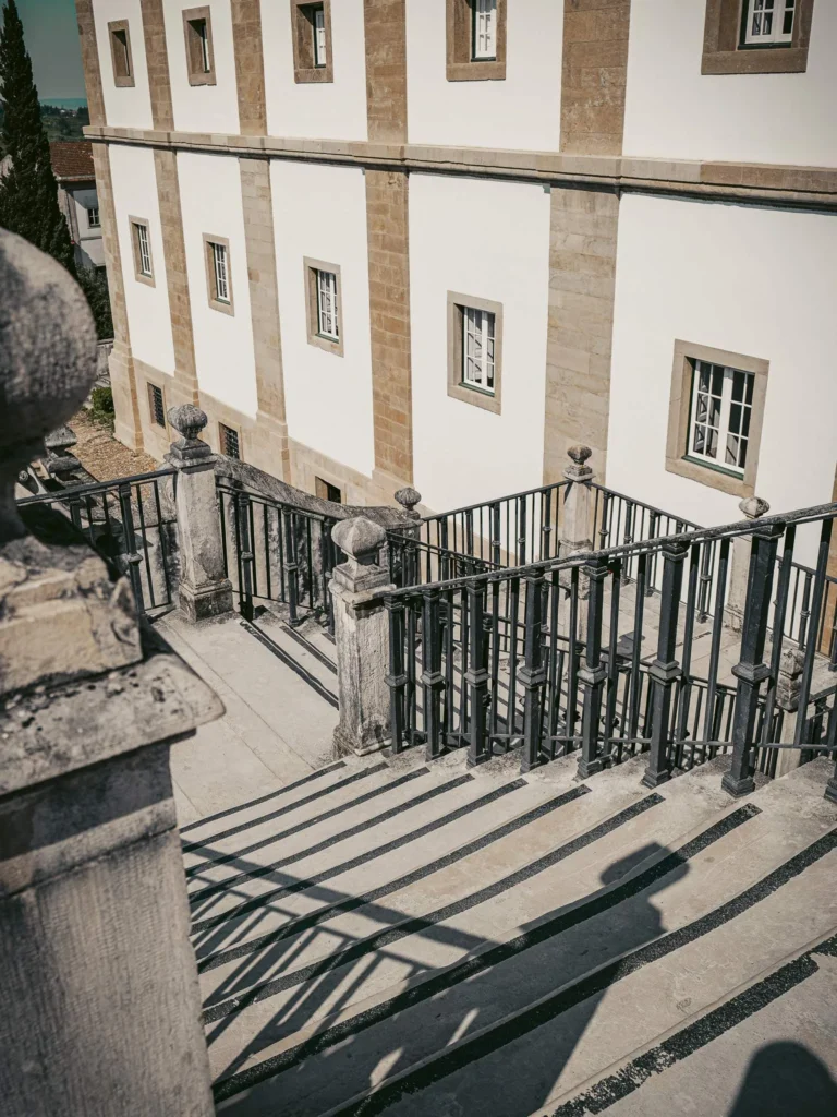 Stone staircase with wrought iron railing, marked shadows and historic building with white facade and stone frames, in Coimbra on a sunny day.