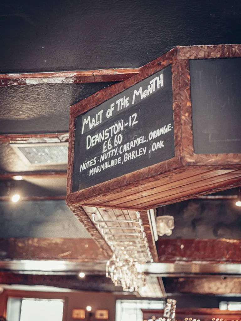 Wooden panel displaying the malt of the month at Ben Nevis Bar in Fort William, with glasses hanging below and a Scottish pub atmosphere.