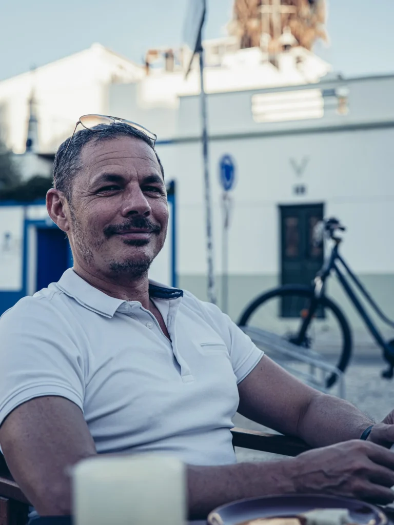 Xavier seated at a terrace table at Bodeguita Salinas in Tavira, tapas and a glass of wine in the summer atmosphere of the Algarve, bike leaning against the white facade.