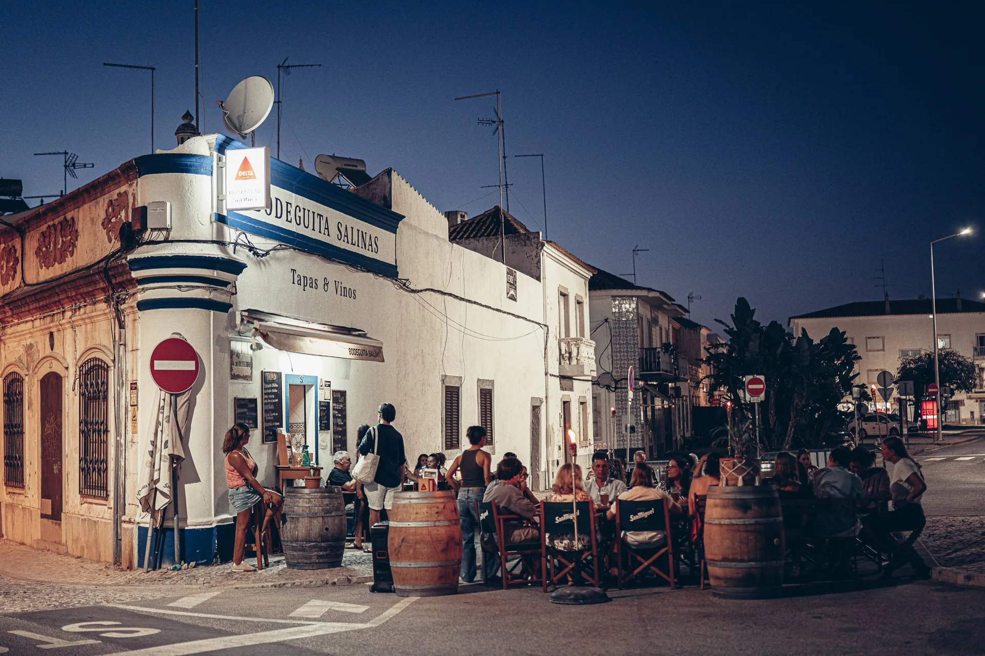 Lively terrace of the Bodeguita Salinas in Tavira on a summer evening: groups seated around wine barrels turned into high tables, sign "Tapas & Vinos," light from the street lamps.
