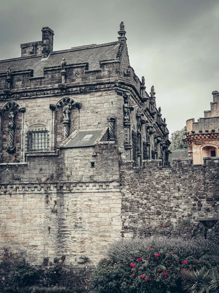 The stone front of Stirling Castle under a cloudy sky, with medieval architectural detail and a flower garden in the foreground.