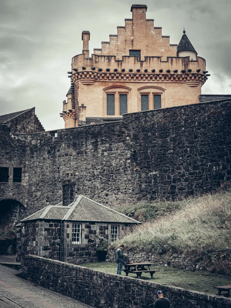 The ramparts and a tower at Stirling Castle, Scotland, under a cloudy sky, with two visitors by a picnic table at the foot of the walls.