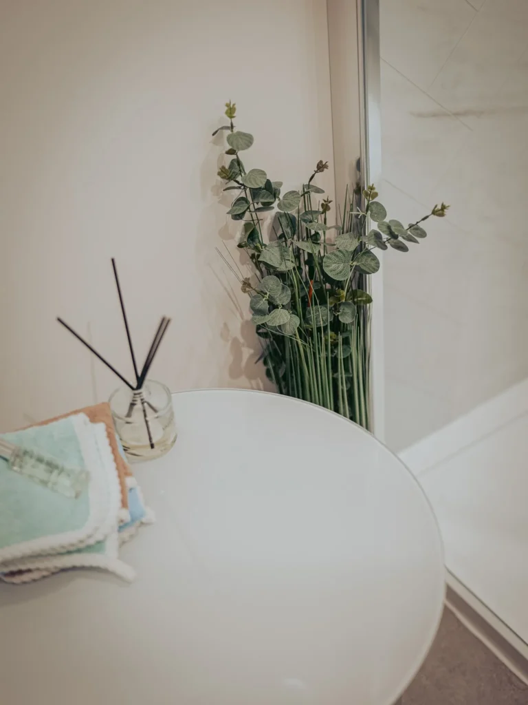 Bathroom corner at Rocklee Bnb in the Highlands with a white table, folded towels, a fragrance diffuser, and a bouquet of foliage near the shower.
