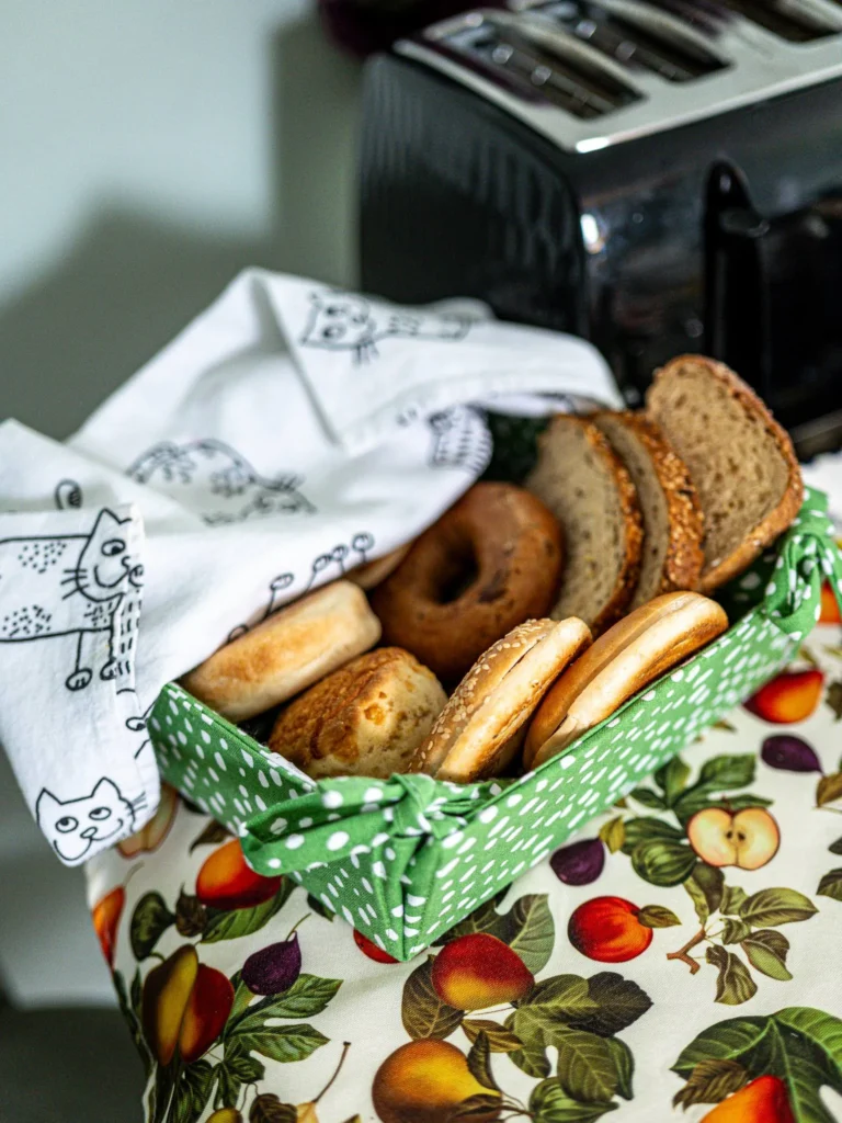 A basket of assorted breads and bagels placed on a fruit-patterned tablecloth, next to a toaster in a kitchen at Rocklee Bnb in the Scottish Highlands in August 2026.