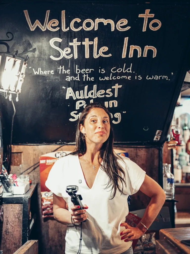 Caroline inside the Settle Inn, in Stirling, in front of a welcome sign, in the dim light of the pub.
