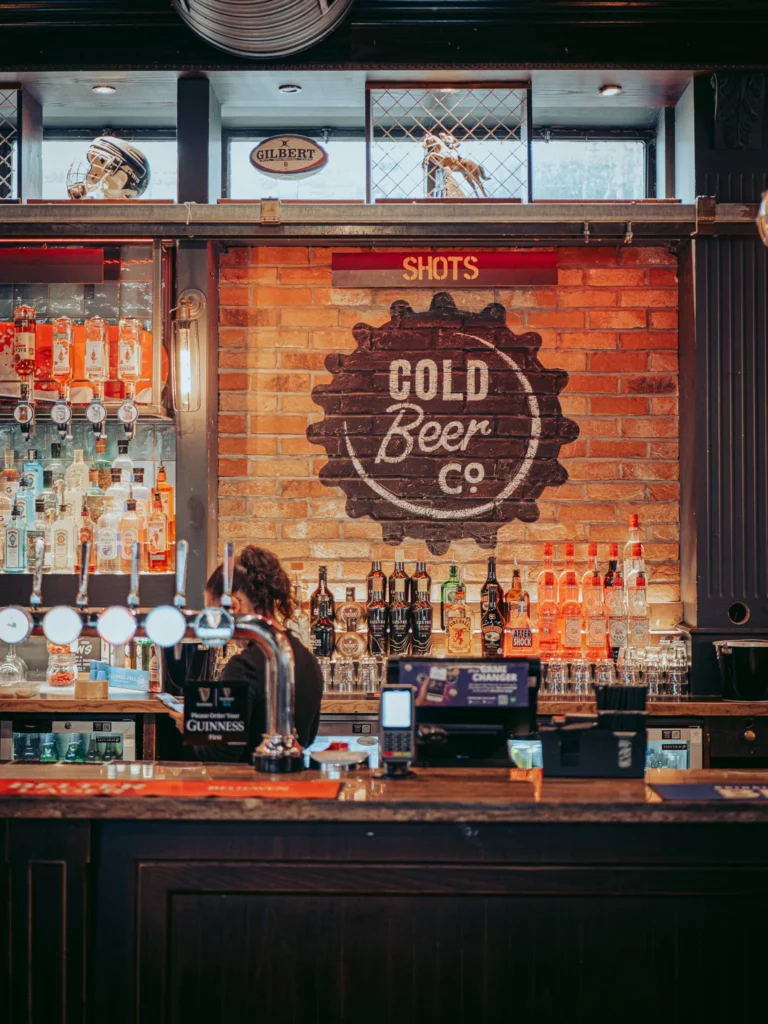 Bar of a pub in Stirling, brick wall adorned with a sign "Cold Beer Co.", a waitress behind the counter.