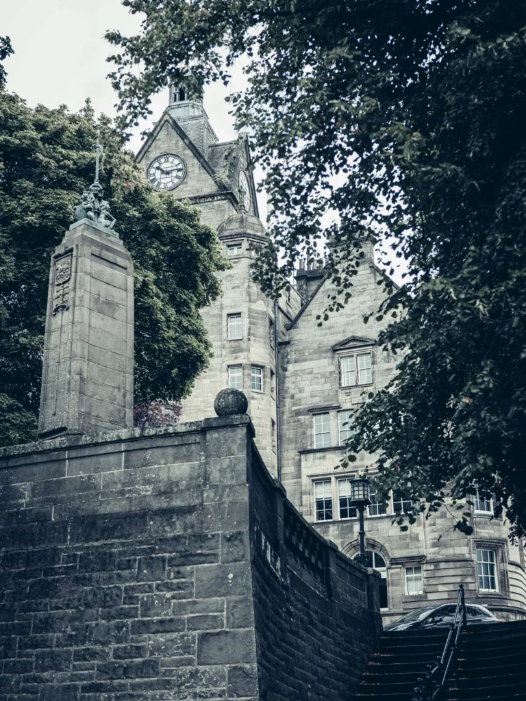 Stone staircase bordered by walls covered in moss, leading up to a large sandstone building with a clock, featuring a black lamppost with a glass lantern. The moss is