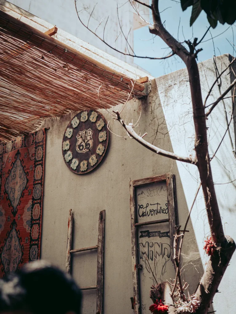 Decorated wall in the semi-covered area of The Garden Bar and Grill in Lagos: carpet hanging on the wall, old wooden ladder, and vintage sign under the thatched roof.
