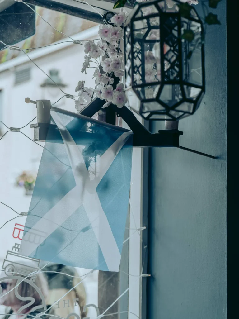 A Saltire stretched against the window of The Geographer in Fort William, with artificial flowers and an iron lantern.