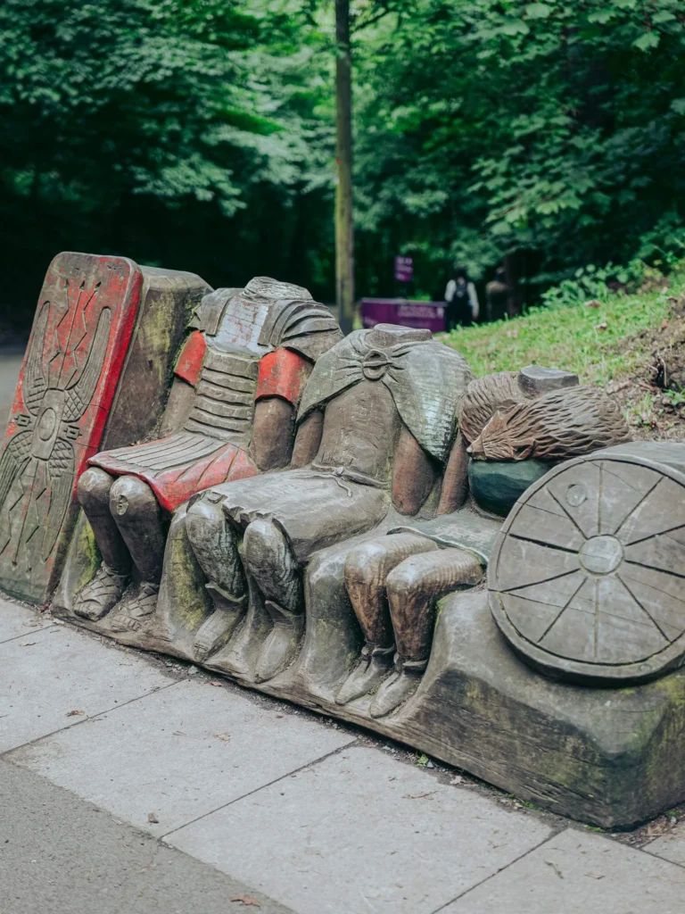 Wooden carving of seated warrior figures without heads, in the woodland beside the Wallace Way near the Wallace Monument in Stirling, Scotland, August 2026.