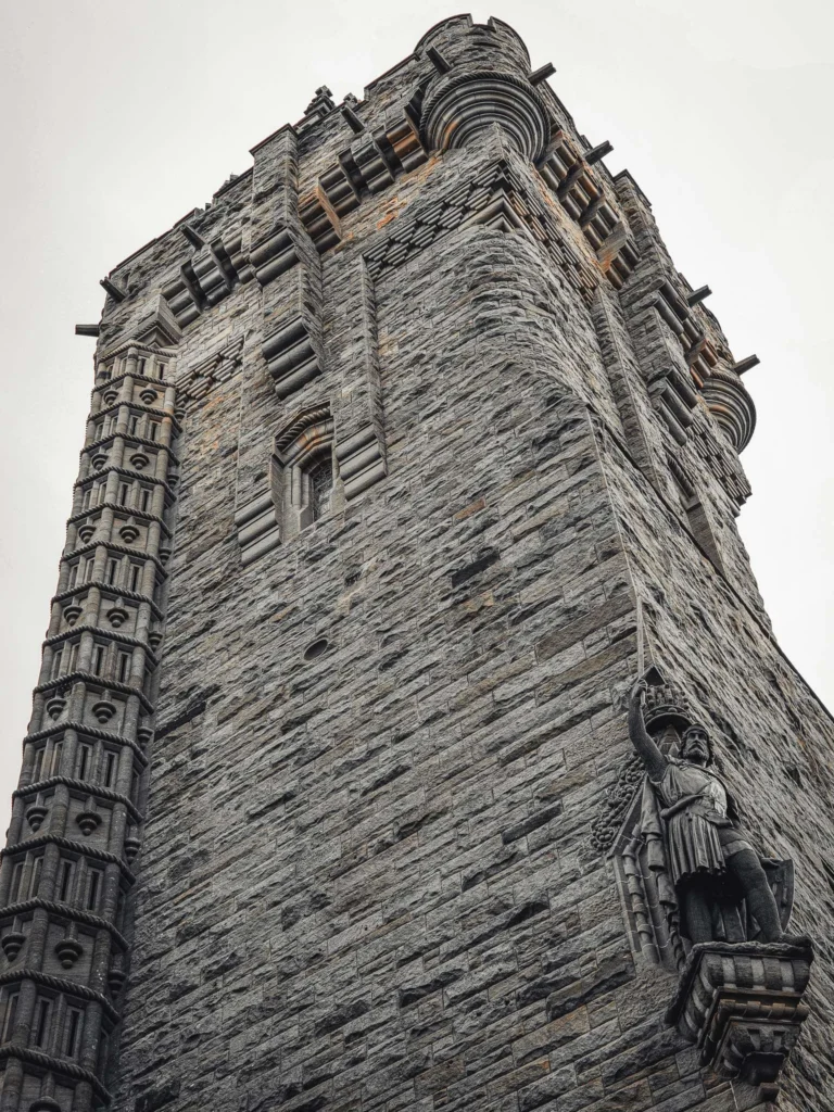 A low-angle view of the Wallace Monument tower in Stirling, Scotland, showcasing the statue of William Wallace and the grey stone architecture under a cloudy sky.