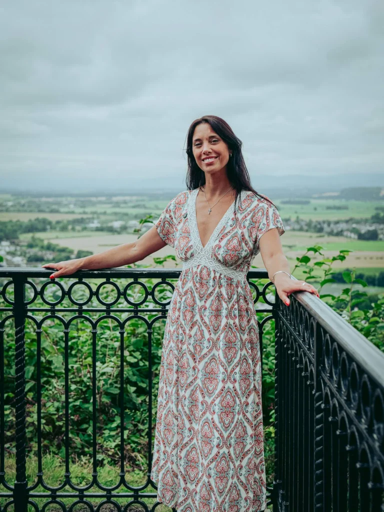 Caroline poses in front of a wrought iron railing with a panoramic view of the lush countryside near the Wallace Monument in Stirling, under a cloudy sky in August 2026.