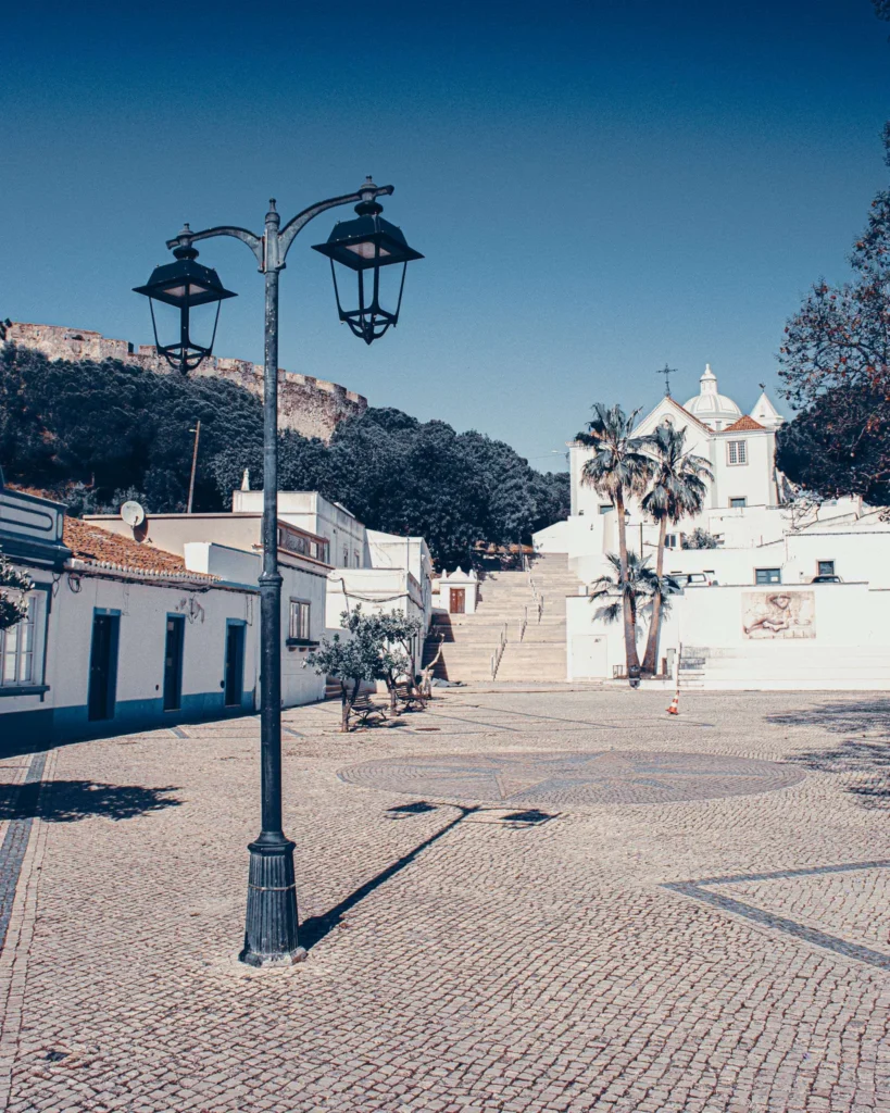 Paved square of Castro Marim with a central lamppost, white houses, stairs leading to a church, and the fortress visible in the background, under a blue sky. Calm and sunny atmosphere in the Algarve, Portugal in May 2023.