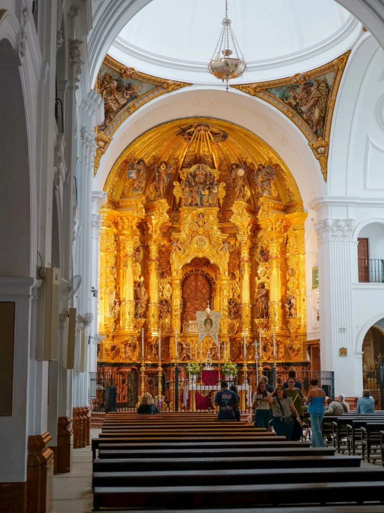 Pala d’altare dorata e interno bianco del santuario del Rocío con visitatori vicino ai banchi